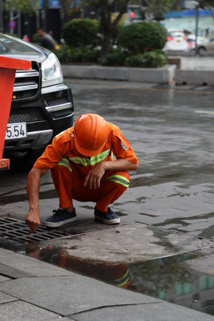 A street worker cleans a drainage system on a wet road after rainfall using a Canon EOS camera.